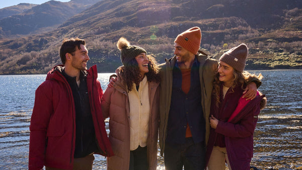 Four friends standing together by a lake with mountains in the background wearin outdoor clothing