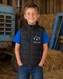 Boy wearing a black padded tractor gilet with blue zips and matching blue T-shirt in a farm barn.