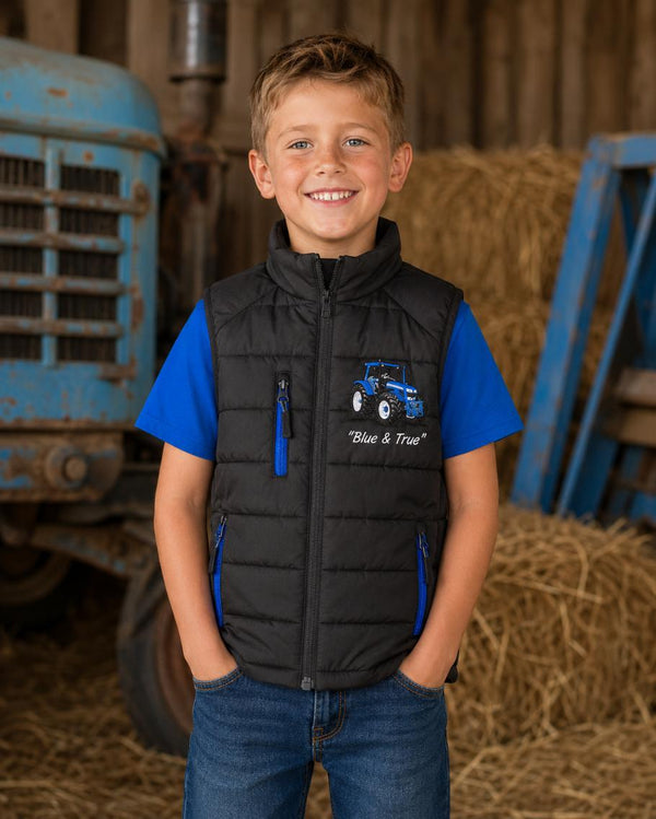 Boy wearing a black padded tractor gilet with blue zips and matching blue T-shirt in a farm barn.