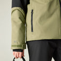 Close-up of a person wearing a technical jacket in olive green and black, showing the sleeve, chest zipper, and hand holding a strap against a light gray background.