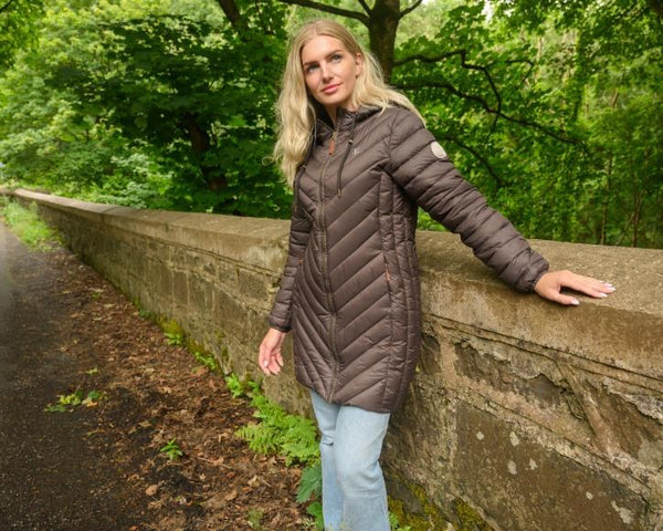 Woman wearing Lighthouse Laurel Cocoa coat outdoors beside a stone wall
