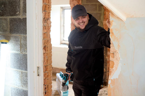 Builder in a black Makita Aero overhead Jacket and cap standing in a room with exposed brick walls.