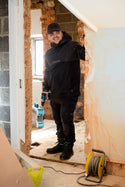 Man in a black jacket standing in a room under renovation with tools and materials around.