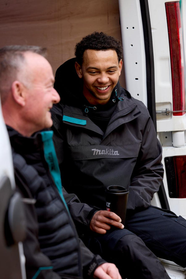 Two men sitting next to a van, one wearing a Makita Aero work jacket.