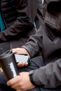 Person wearing a Makita waterproof overhead jacket holding a smartphone and a black cup.