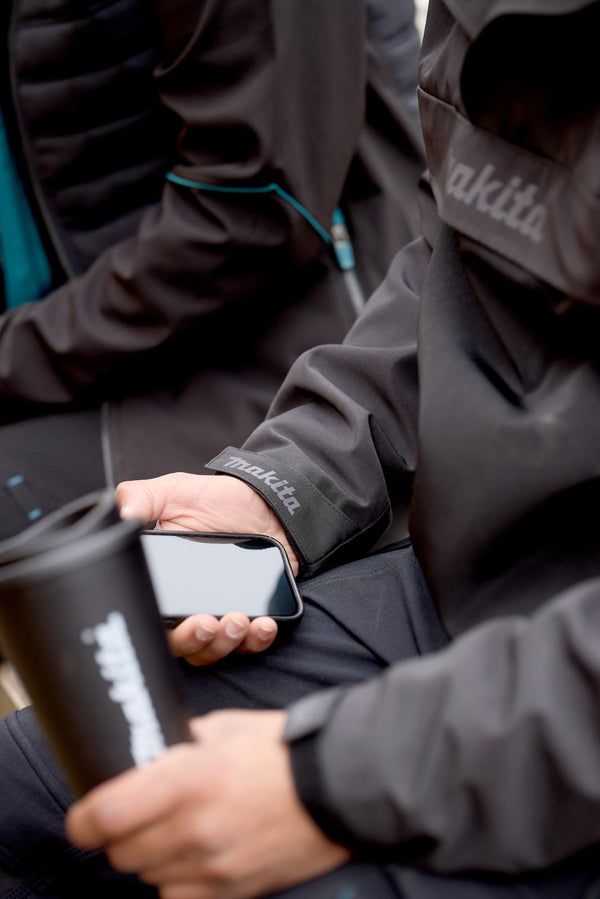 Person wearing a Makita waterproof overhead jacket holding a smartphone and a black cup.