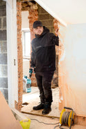 Person in a black jacket holding a drill in a room under construction.