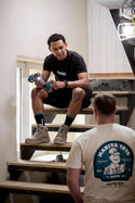 Two men sitting on a staircase, one wearing a black shirt with a brand logo and the other wearing a white shirt with a blue logo.