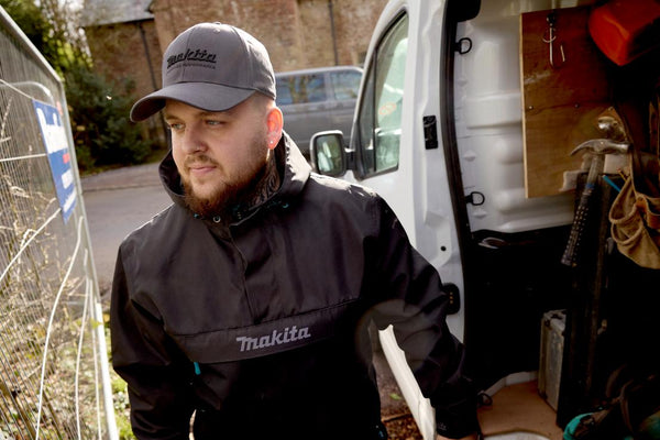 builder wearing a Makita cap and jacket standing next to a van with tools inside.