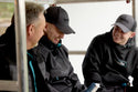 Three men wearing Makita black jackets, hoodies and hats, sitting together on a vehicle.