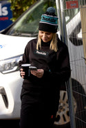 Woman wearing a Makita womens work hoodie and work beanie, holding a cup and phone, standing next to a car.