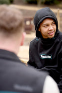 Person wearing a black hoodie with Makita logo interacting with another person outdoors.