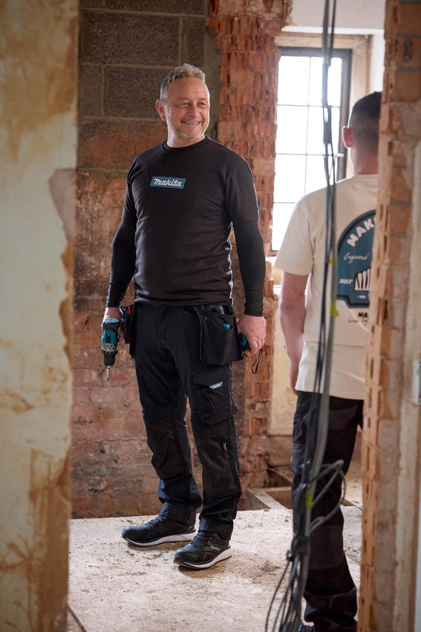 Two men in a room with exposed brick walls and a window, one wearing a black Makita shirt.