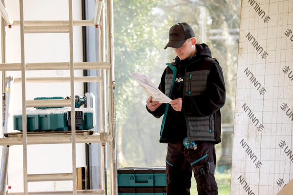 Builder in Makita work attire reading a document next to a ladder and tools indoors.