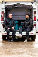 Two men sitting in the back of a van with tools, on a wet ground.