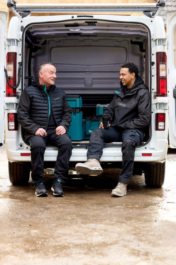 Two men sitting in the back of a van with tools, on a wet ground.