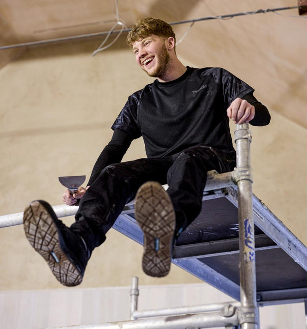Man sitting on a metal structure wearing black  Makita workwear.