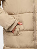 Close-up of a beige puffer jacket with horizontal quilting, a ribbed sleeve cuff, and a hand reaching into a front pocket.