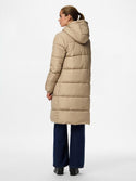 A woman shown from the back wearing a long, light beige puffer coat with a hood, paired with dark blue wide-leg jeans and black shoes, against a plain white studio background.