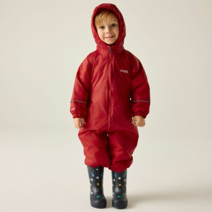 A toddler wearing a bright red hooded snowsuit with a front zipper and elastic cuffs, paired with dark rain boots featuring a colorful space print, standing against a plain light beige studio background.